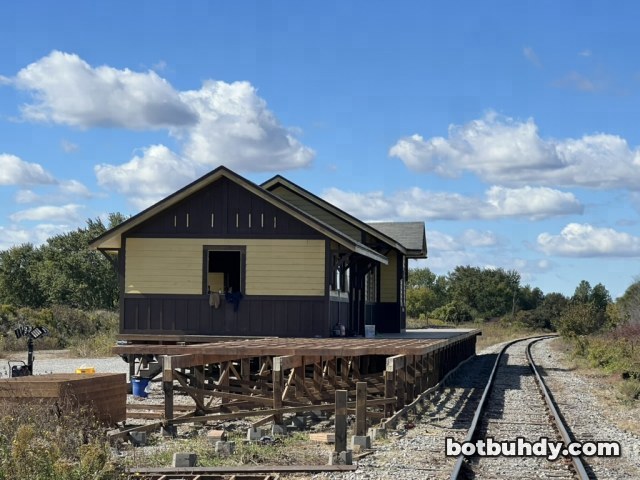 The station set and its wooden platform beside the railway tracks under a cloudy blue sky.