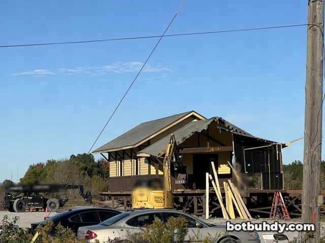 A yellow excavator beside the station porch, with parked cars in front.