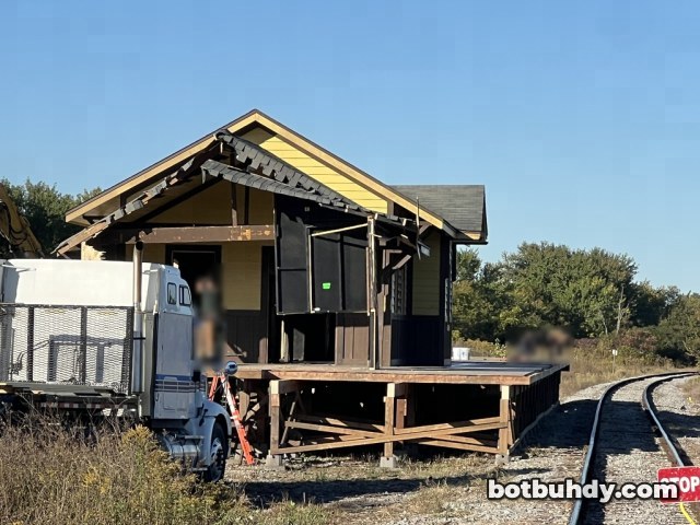 The station by the tracks with its porch roof hanging down, a white truck at left and a STOP sign in front.