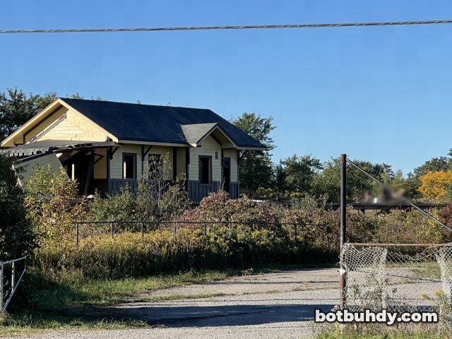 The station seen past tall weeds and a fence, with its porch roof collapsed.
