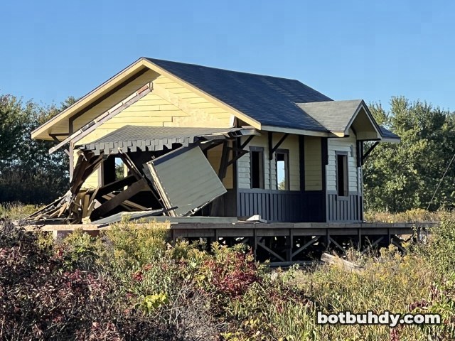 Close view of the station front with a wall panel hanging loose over splintered porch boards.