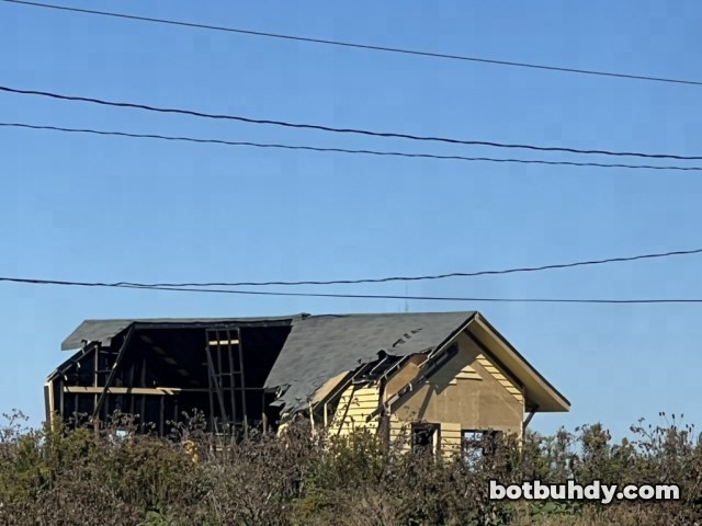 The station seen behind weeds and power lines, with one half of its roof torn away and the frame exposed.