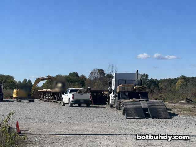 A gravel lot with a white pickup, trucks and an excavator beside an empty wooden platform frame.