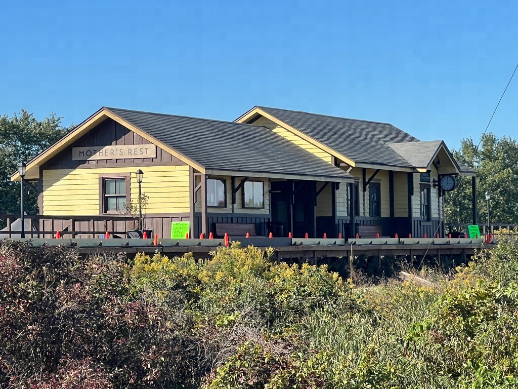 The yellow Mother's Rest station set seen across green and yellow brush, with orange safety cones along the deck and a round clock on the building