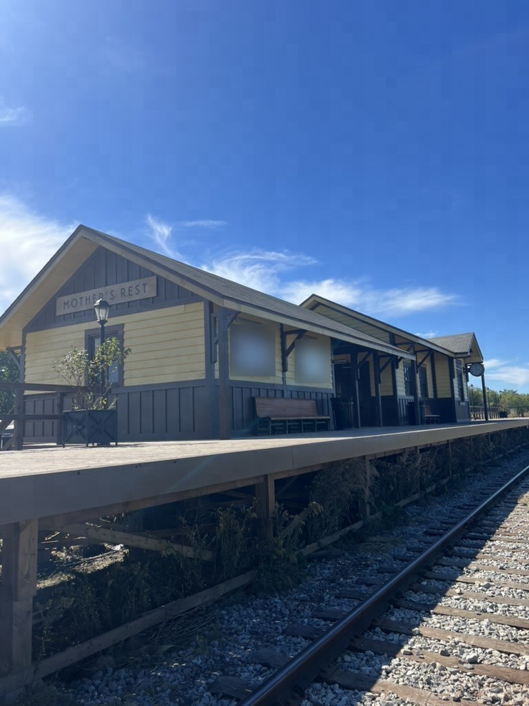 The long wooden platform of the Mother's Rest station set in the midday sun, with the MOTHER'S REST sign, a lamp post, a bench and the rails running alongside