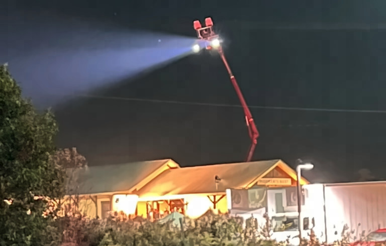 A red boom lift raises a bright light that beams across the night sky above the lit Mother's Rest station roof.