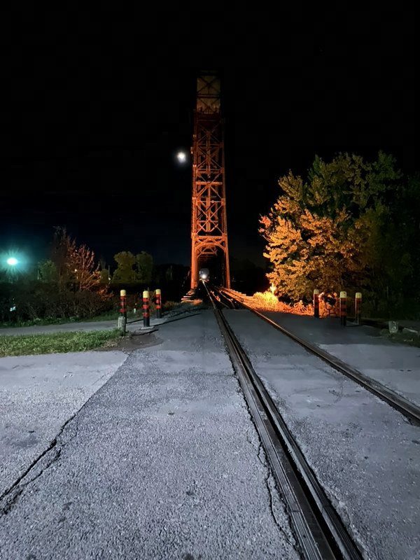 Railway tracks run straight across pavement toward an orange-lit bridge tower at night, with a lit tree and a bright moon.