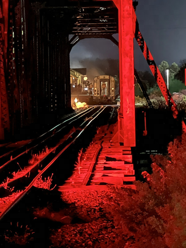 Red-lit steel bridge beams frame the rails and, far beyond, the back of a train beside the glowing station.
