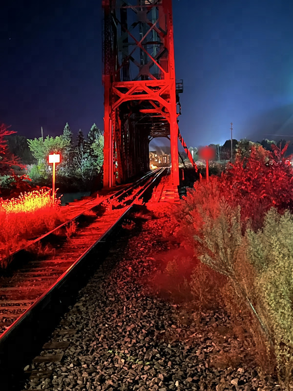 Railway tracks bathed in red light lead under a tall steel bridge tower at dusk, with the lit station small in the distance.