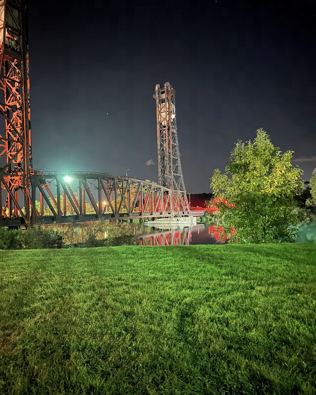Two tall steel bridge towers and the span between them at night, seen across a green lawn.