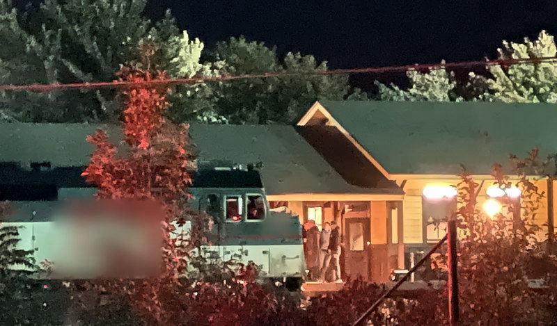 At night, a green-and-white locomotive stands beside a lit station porch where a few crew members gather, with red-leaved shrubs in front.