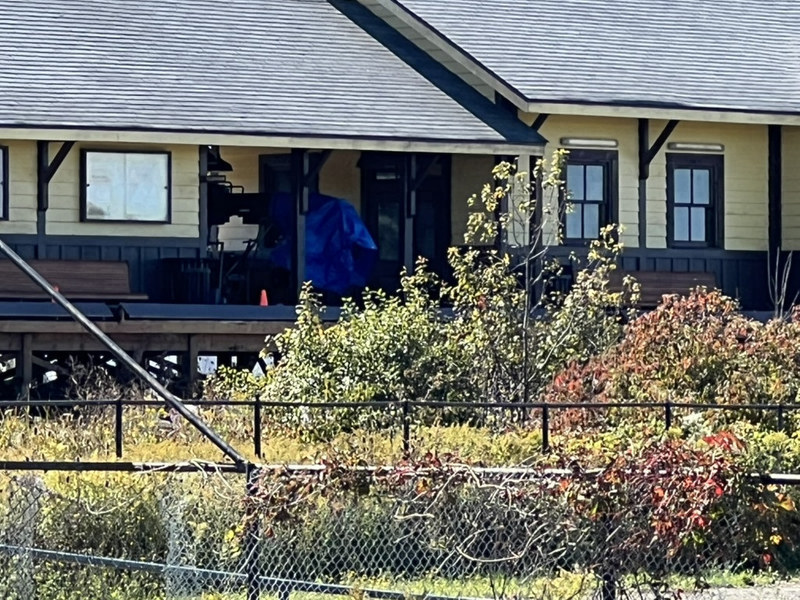 The station porch in daylight with a rig under a blue tarp, benches and an orange cone, seen over a fence and autumn shrubs.