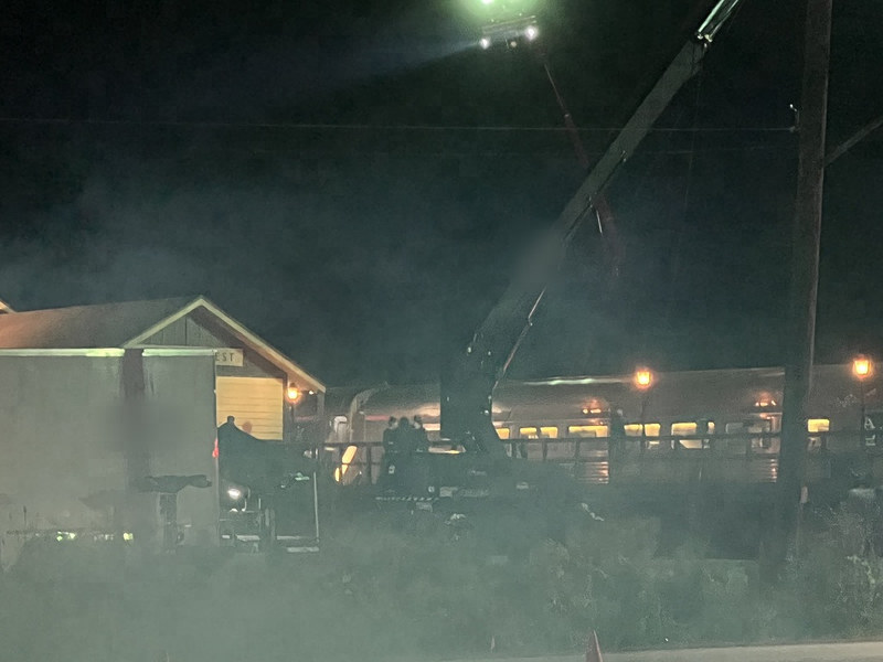 A tall crane with a bright lighting head over the station set at night, haze drifting across the platform and the parked train.