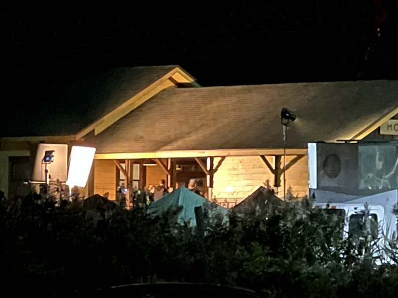 The station porch glowing under film lights at night, with light panels at left and a tent among the shrubs.