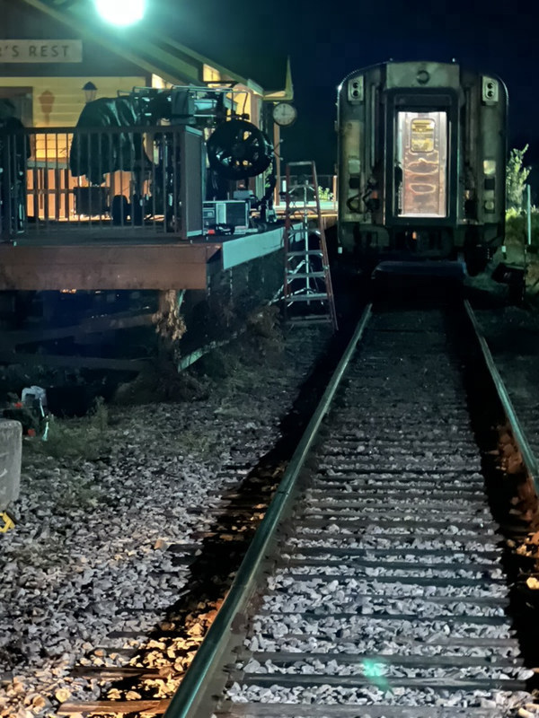 Looking along the rails at night to a parked passenger car beside the station deck, where camera and lighting gear stand by a ladder.