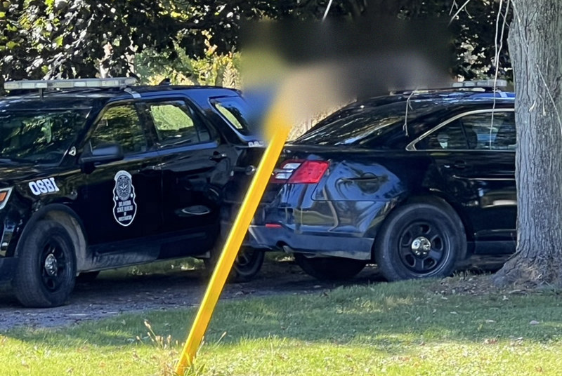A black prop SUV marked OSBI parked beside a dark unmarked sedan with a light bar, on grass under trees.