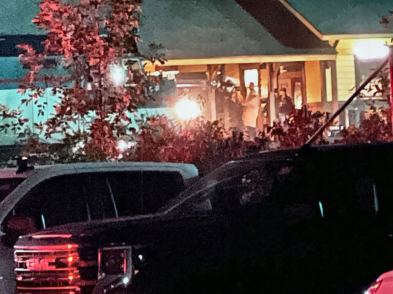 Night view of the lit station deck with several crew members and lights gathered around a man, seen past a red-leaved tree and parked vehicles.