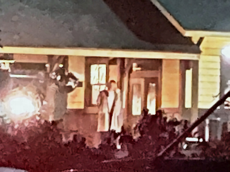 Enlarged close view of the lit porch with a man in a long light coat standing near the doorway.