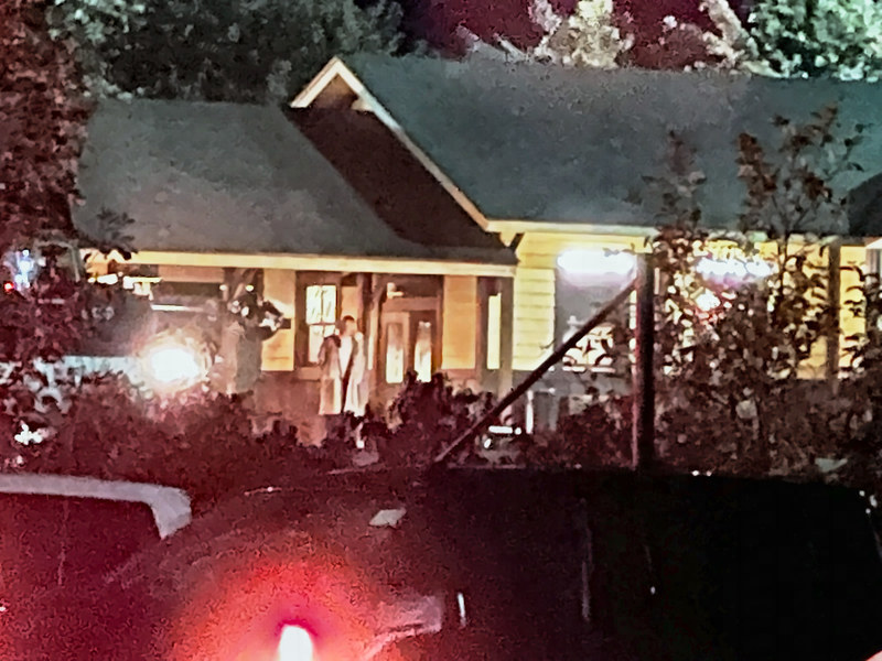 A man in a long light coat stands on the lit station deck at night, seen past a red-leaved tree and parked vehicles.