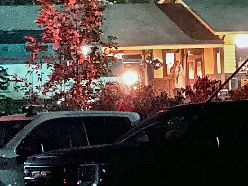 Night view past parked vehicles and a red-leaved tree to the lit porch, with a train's nose at left and a man in a long light coat on the deck.