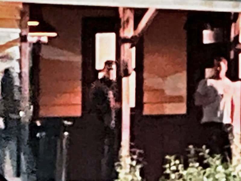 Enlarged close view of the porch: a man in a white T-shirt at the doorway and a person in dark clothes at left.
