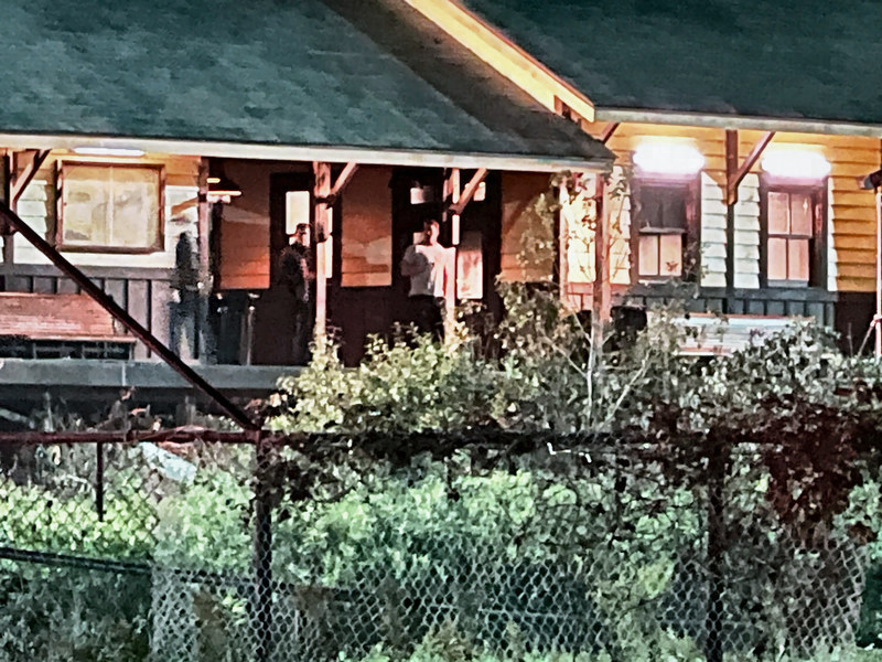 The lit station porch at night seen over a chain-link fence and shrubs, with a man in a white T-shirt by the doorway and two people in dark clothes nearby.