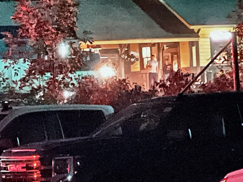 Night view past a red-leaved tree and parked vehicles to the lit deck, where a man in a white T-shirt stands near a camera rig.