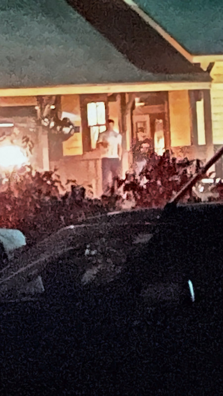 Tall enlarged crop of the lit porch with a man in a white T-shirt standing on the deck.