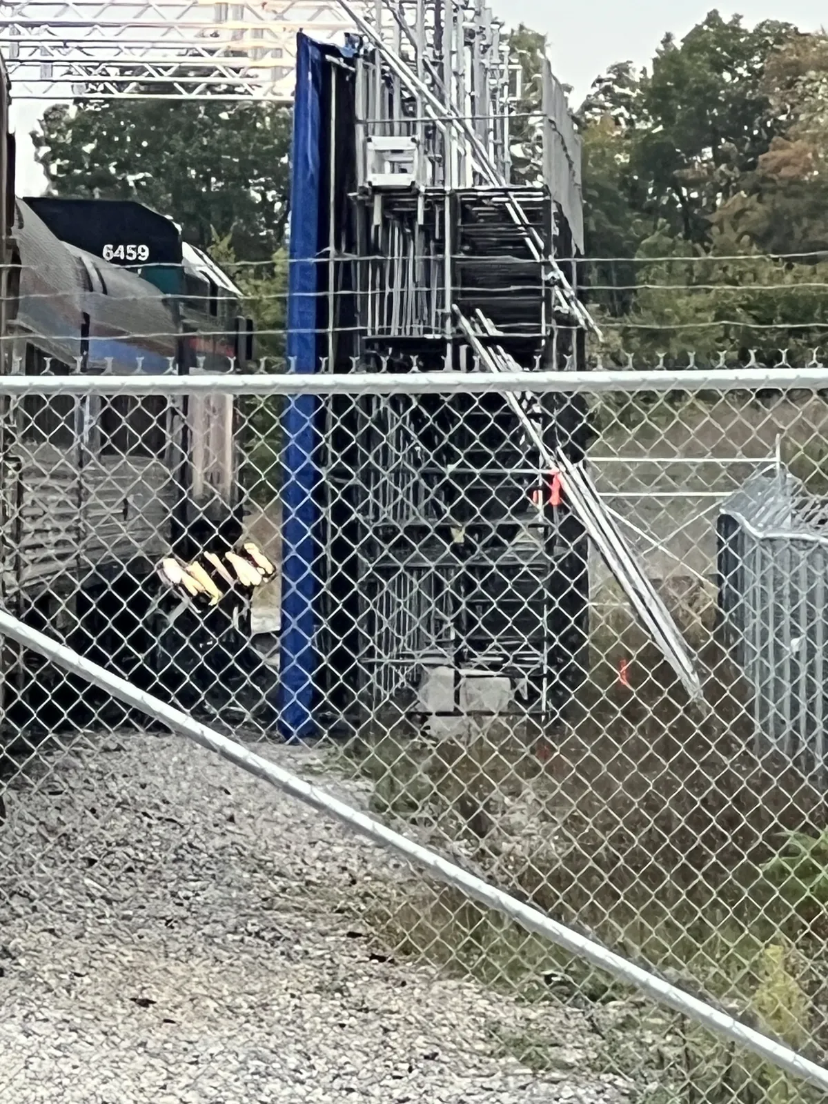 Seen through a fence: the back of the scaffold tower, with the blue screen hanging on the side facing the train and a truss grid overhead