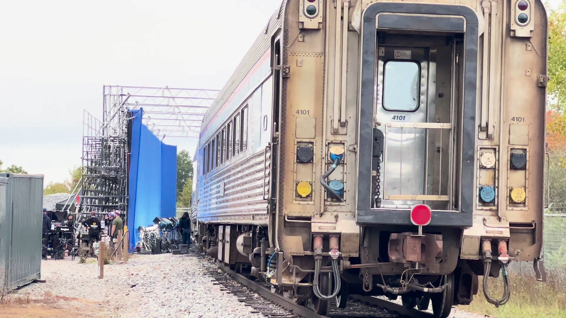 A stainless-steel passenger car on track. To its left, a tall scaffold tower holds a blue screen, with a truss grid overhead and a row of film lights on stands at the base.