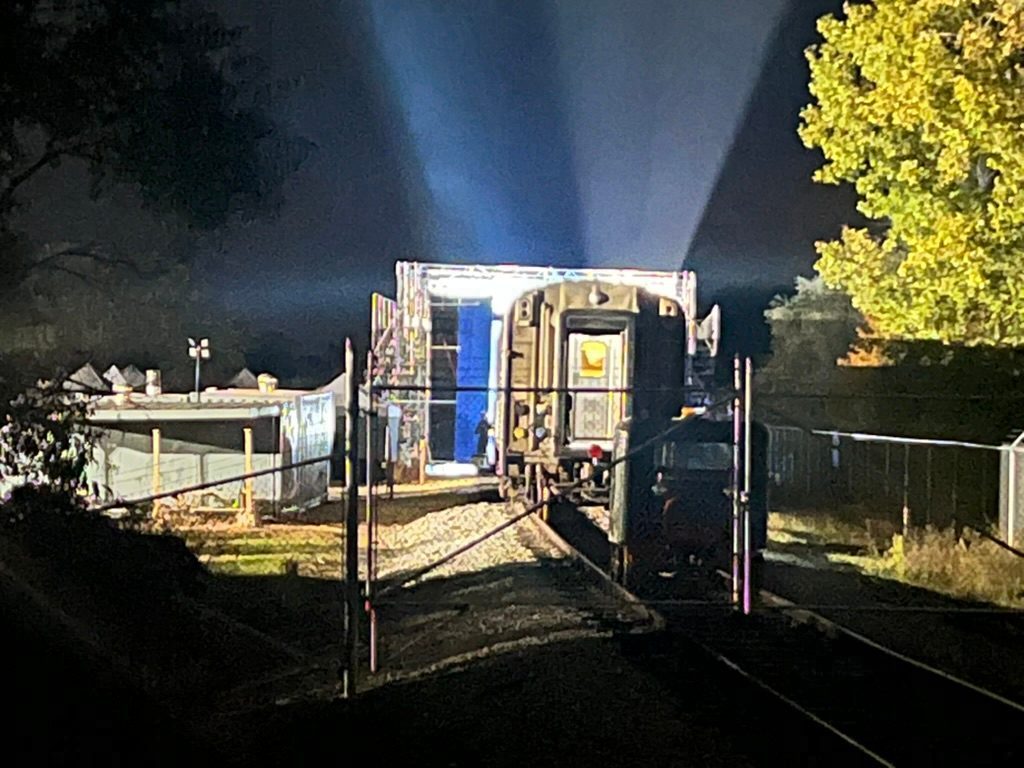 At night, the end of the parked passenger car on its track, with the lit scaffold tower and a strip of blue screen beside it and beams of light shining up into the dark sky