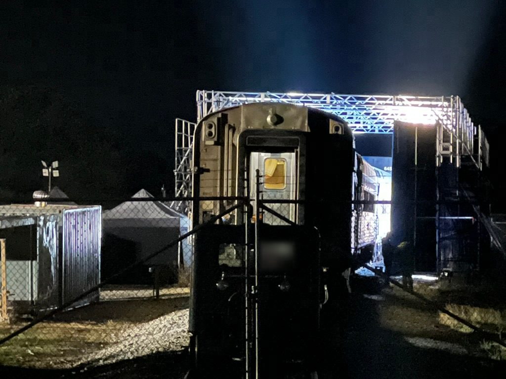At night, seen through a fence: the end of the parked passenger car under the brightly lit truss grid, with light glowing along the screen side of the car